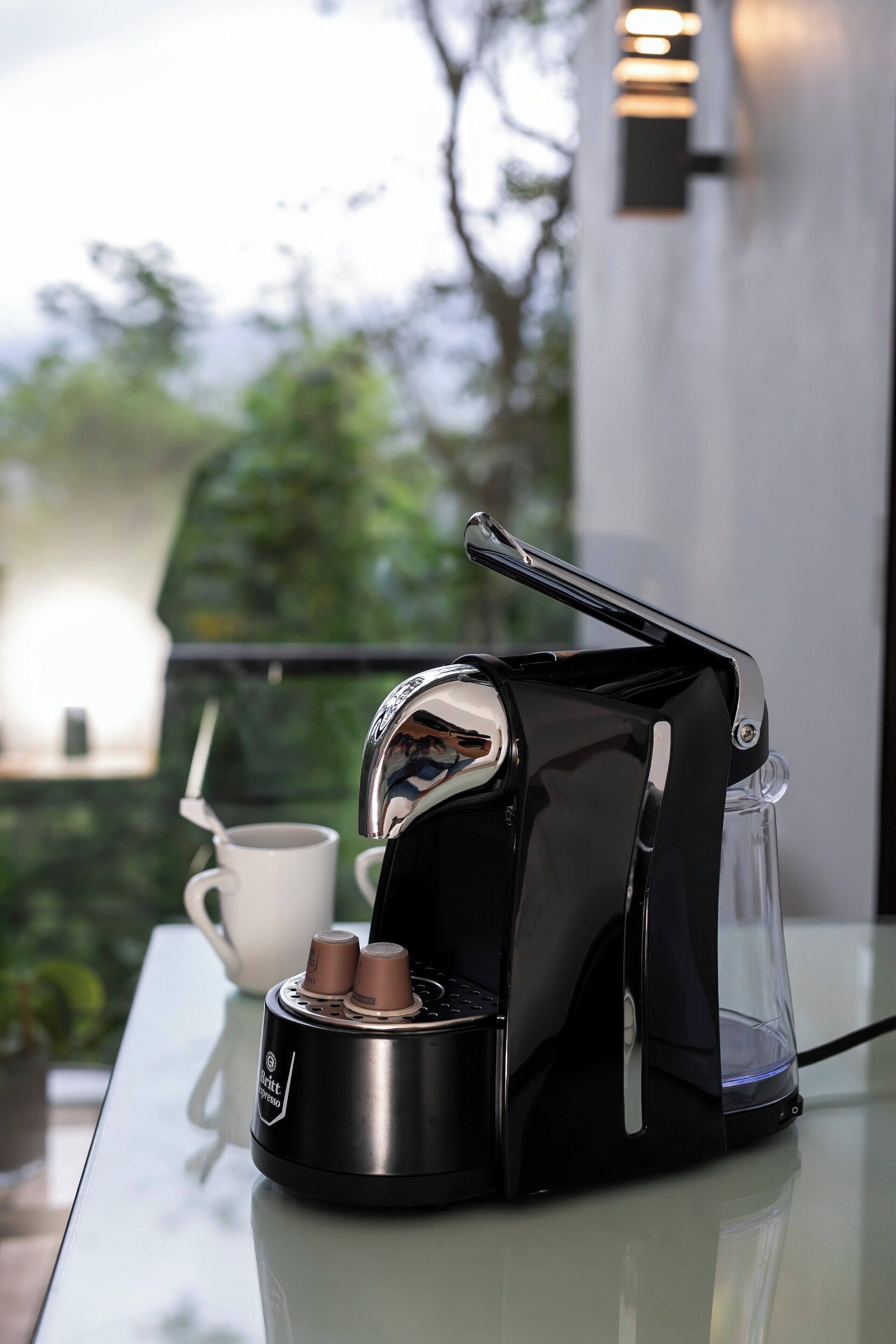 Une machine à café est posée sur une table en verre, à côté se trouve une tasse blanche, une verseuse en verre transparent et une cuillère.