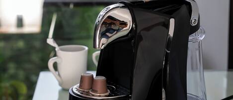 Une machine à café est posée sur une table en verre, à côté se trouve une tasse blanche, une verseuse en verre transparent et une cuillère.
