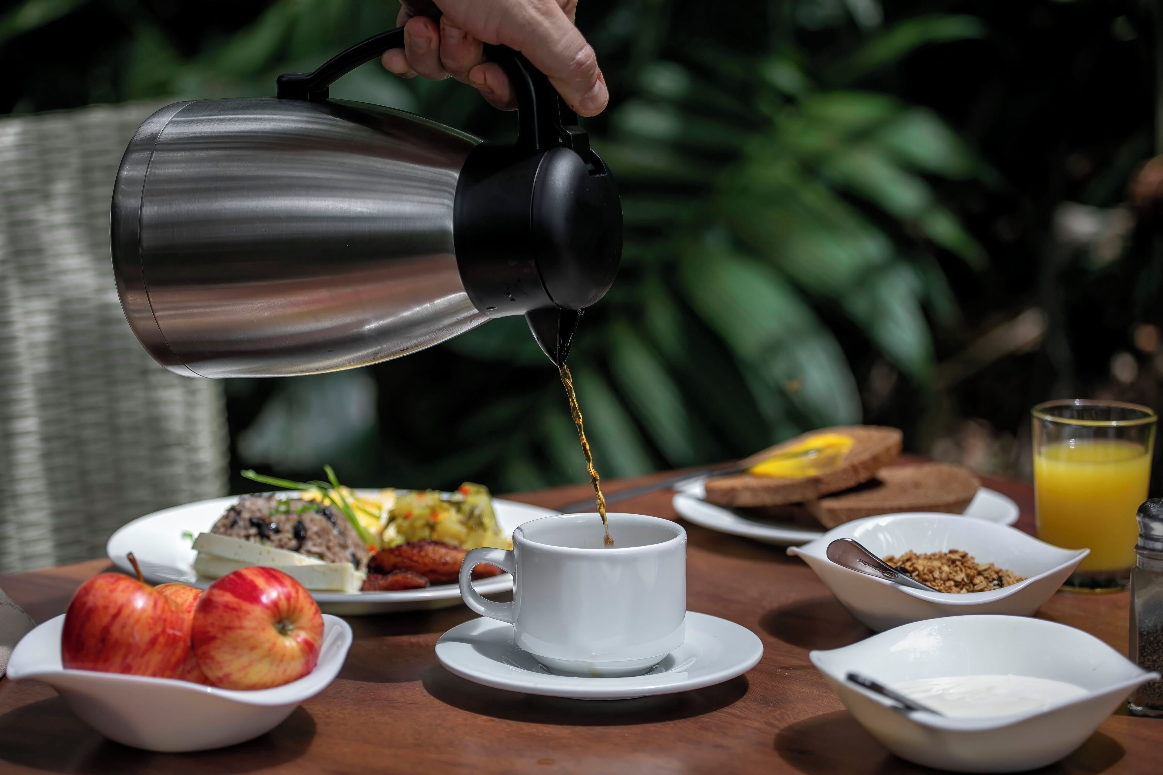 A breakfast setting with a coffee pot pouring into a cup, surrounded by plates of food and a glass of orange juice.