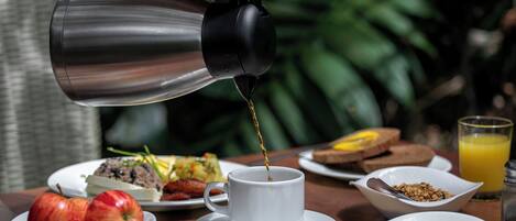 Une table mise pour le petit-déjeuner, avec une cafetière versant du café dans une tasse, entourée d’assiettes remplies de nourriture et d’un verre de jus d’orange.