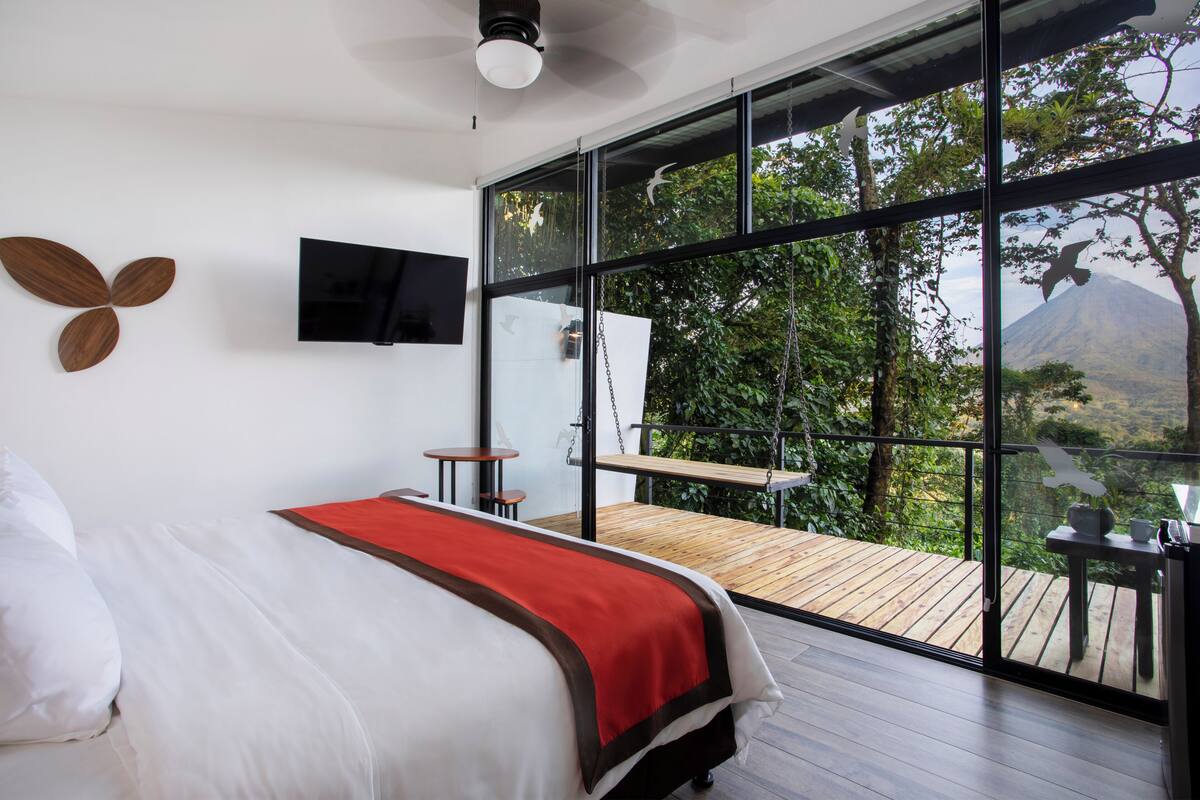 Ein modernes Schlafzimmer mit einem großen Bett, einem an der Wand befestigten Fernseher und einem Balkon mit Blick auf Bäume und ein Bergpanorama.