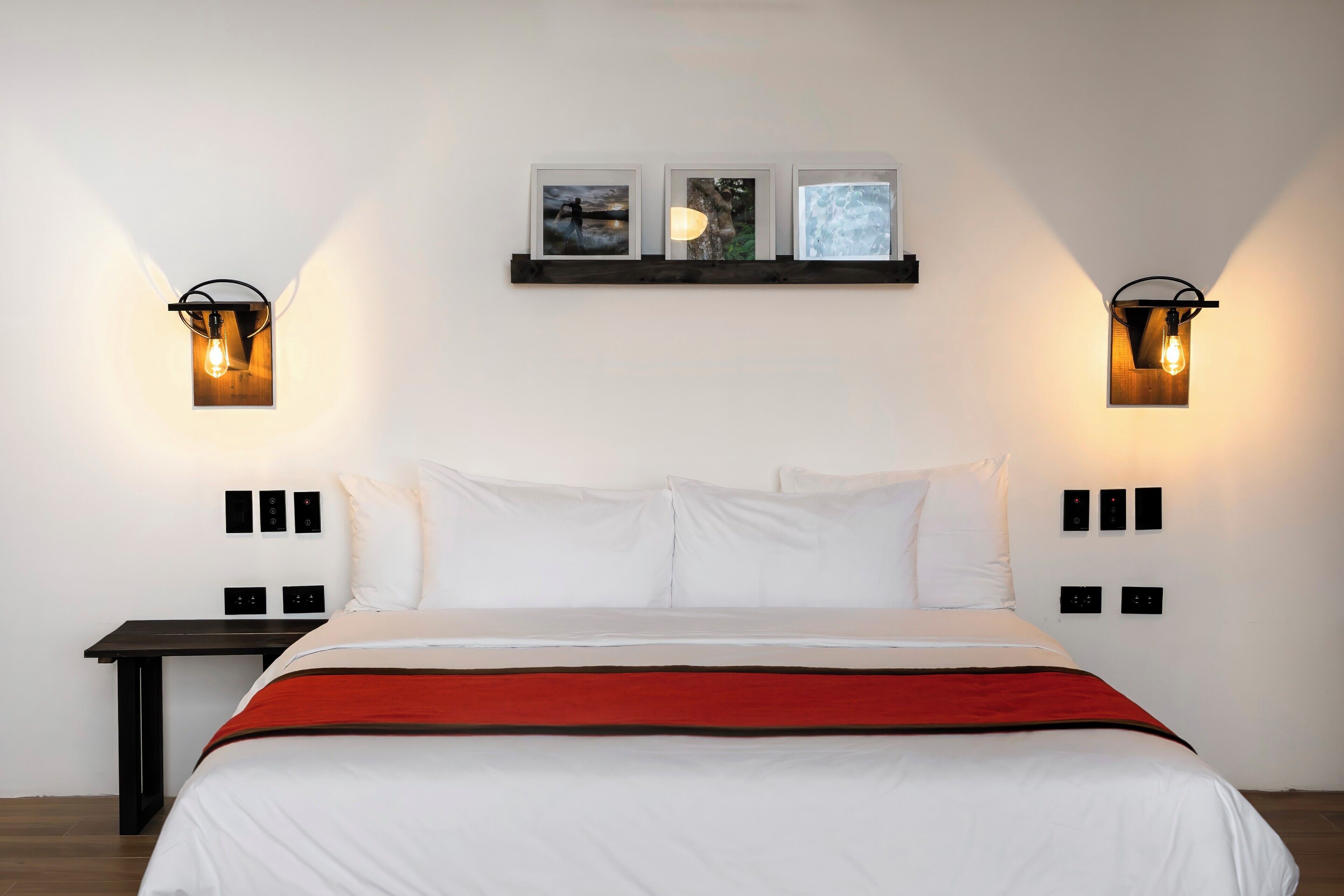 Una cama bien hecha con ropa de cama blanca y roja, dos lámparas fijadas a la pared y tres cuadros colgados sobre la cama.