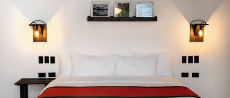Una cama bien hecha con ropa de cama blanca y roja, dos lámparas fijadas a la pared y tres cuadros colgados sobre la cama.