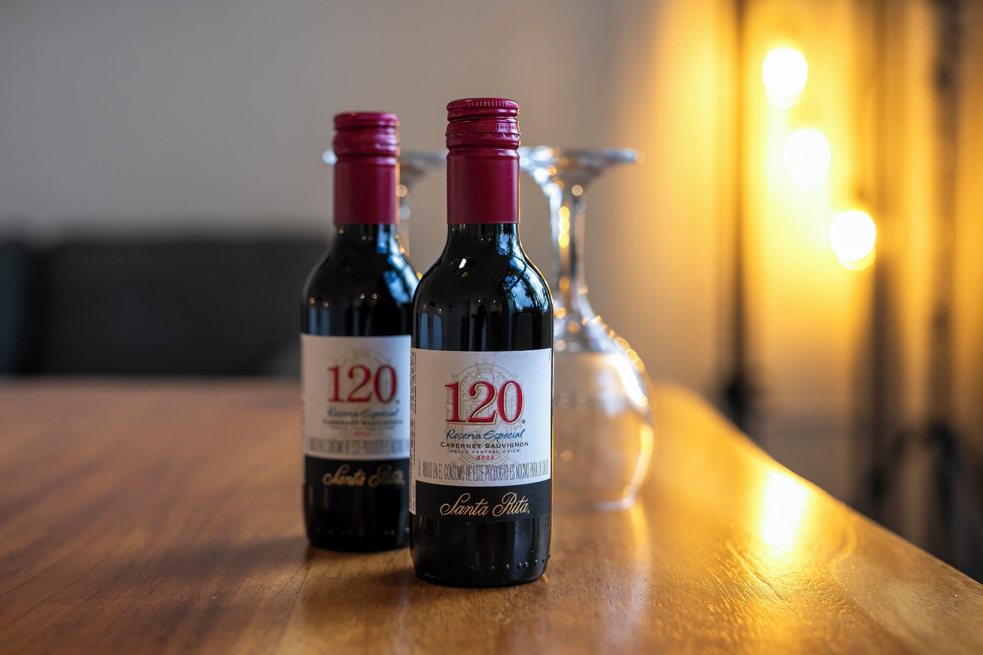 Two bottles of wine with red caps on a wooden table, with a glass and a blurred background.