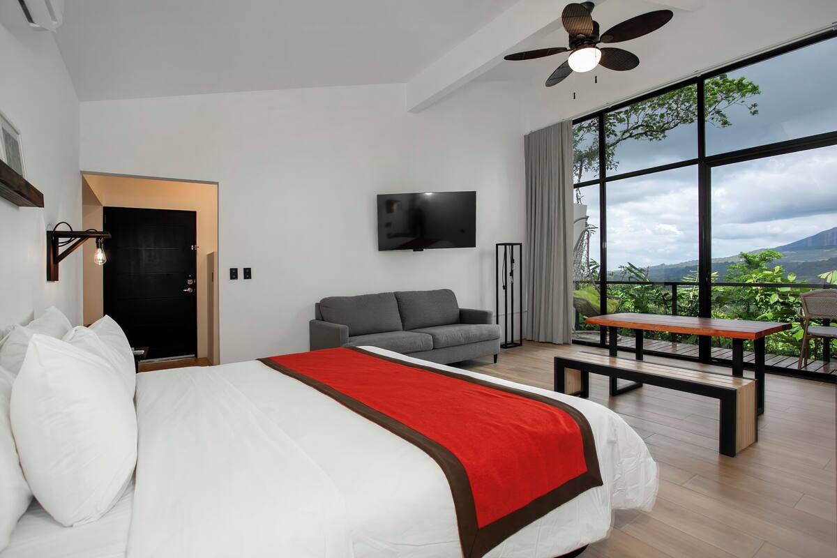 Ein modernes Hotelzimmer mit einem großen Bett, einem grauen Sofa, einem an der Wand montierten Fernseher und einem großen Fenster mit Blick ins Freie.