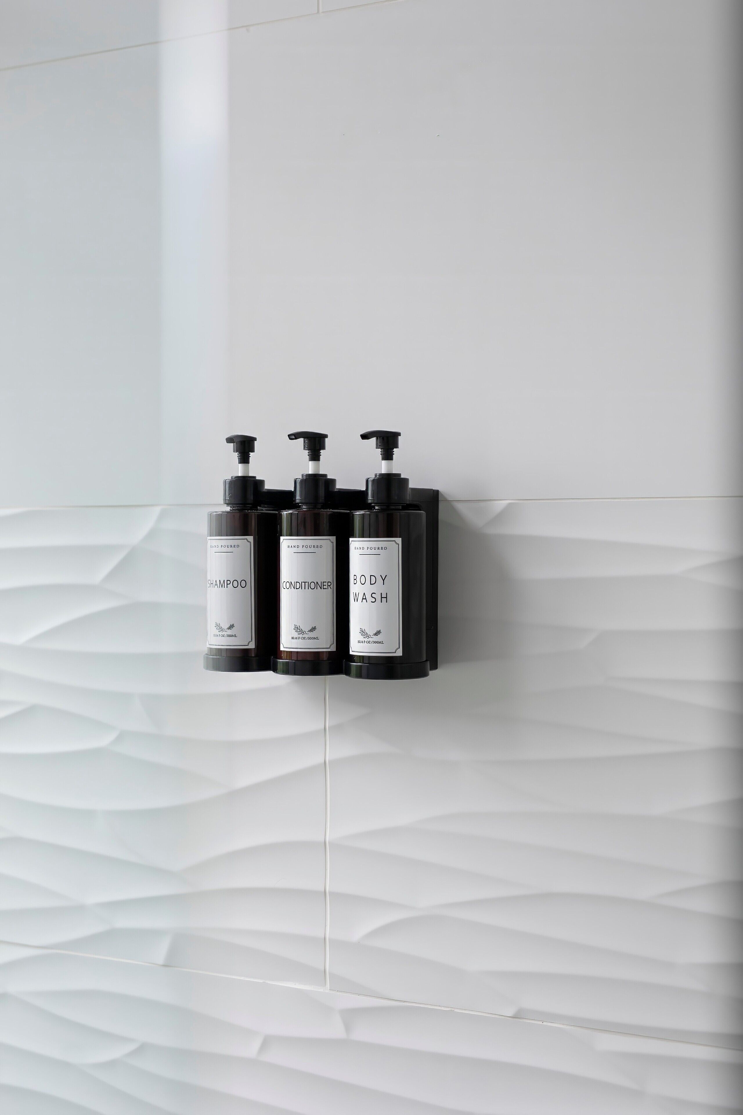 Three black bottles of shampoo, conditioner, and body wash mounted on a white textured wall.