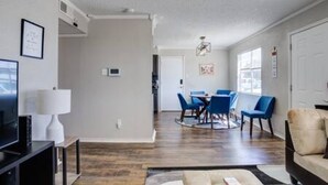 Uma sala moderna com televisão de tela plana, uma área de jantar com cadeiras azuis e um corredor que leva a uma porta.