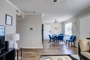 A modern living room with a flat-screen TV, a dining area with blue chairs, and a hallway leading to a door.