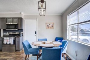 A modern kitchen with stainless steel appliances, a dining area with blue chairs, and a large window with blinds.