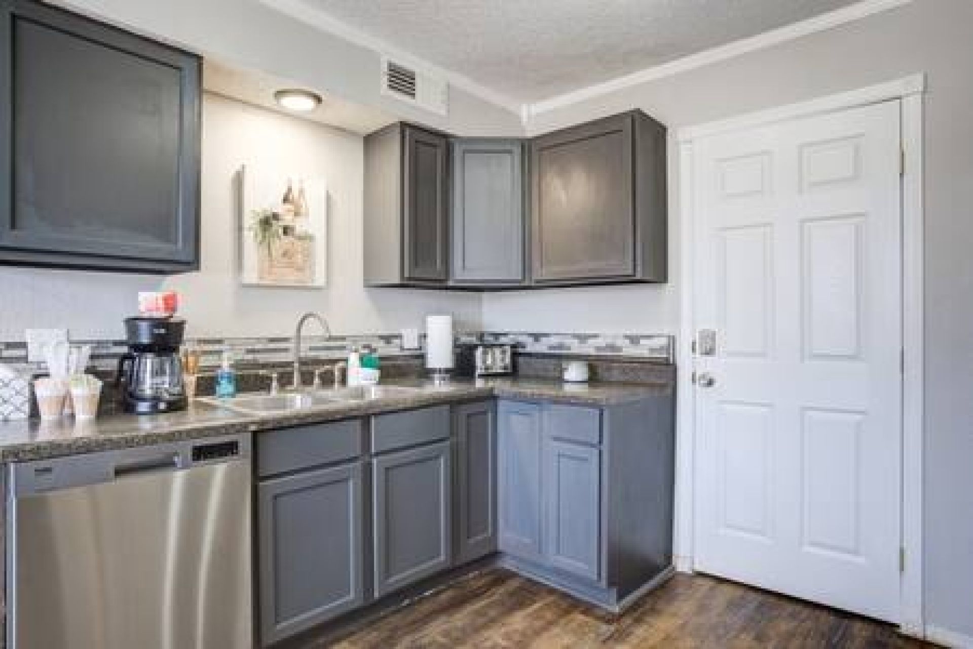 A kitchen with grey cabinets, a dishwasher, a coffee maker, and a white door.