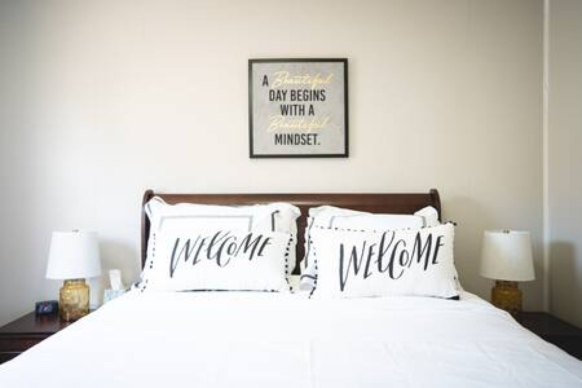 A neatly made bed with two pillows, each with the word 'WELCOME' on them, and a framed inspirational quote on the wall above.
