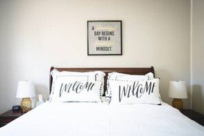 A neatly made bed with two pillows, each with the word 'WELCOME' on them, and a framed inspirational quote on the wall above.