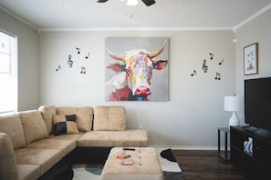Un salon avec un canapé d’angle beige, une table basse, un tableau représentant une vache avec des notes de musique et un téléviseur posé sur un meuble.