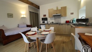 A compact kitchen with wooden cabinets, a microwave, a gas stove, and a sink. There is a dining table with chairs and a small sofa in the adjacent living area. - Gîte St Michel - SAINT-CLEMENT-LES-PLACES (SAINT-CLEMENT-LES-PLACES)