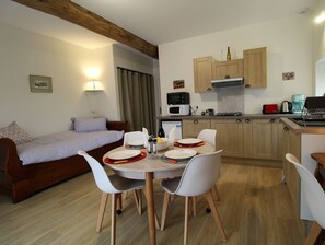 Een compacte keuken met houten kastjes, een magnetron, een gasfornuis en een spoelbak. In de aangrenzende woonkamer staat een eettafel met stoelen en een kleine bank.