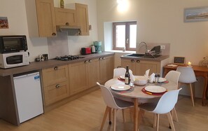 Een compacte keuken met houten kastjes, een magnetron, een gasfornuis en een spoelbak. Er is een eettafel met stoelen en een kleine koelkast.
