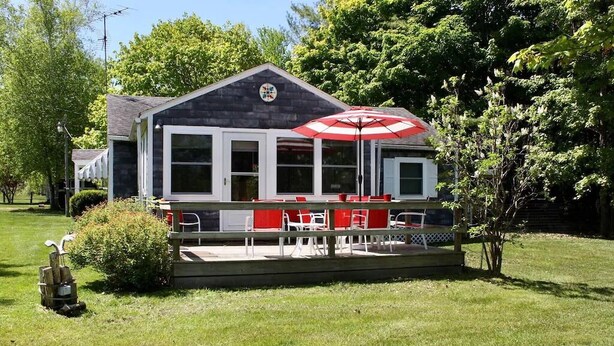 A building with a covered patio area, featuring outdoor seating and a red umbrella. - In the narrows on Lake Leelanau, pet friendly, sleeps 8, and a dock! (Lake Leelanau)