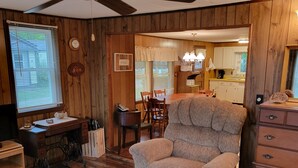 A cozy living room with a wooden floor, a large armchair, a sewing machine, and a dining area with a table and chairs. - In the narrows on Lake Leelanau, pet friendly, sleeps 8, and a dock! (Lake Leelanau)