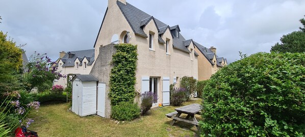 A house with a steeply pitched roof, a small garden with a wooden bench, and a shed.
