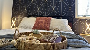 A bedroom with a geometric patterned headboard, a bed with white and brown bedding, a bedside table with a lamp, and a window with a view of trees.