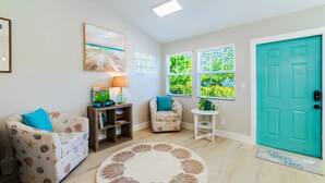 A living room with a teal door, a round rug, two armchairs, a bookshelf, a lamp, and a window with greenery outside.