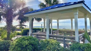 A covered pavilion with a view of the ocean, surrounded by palm trees and greenery. - Stylish Oceanfront Condo - Huge Balcony - 3 Pools, Lazy River, and Hot Tub (Myrtle Beach)