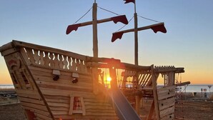 A wooden pirate ship play structure with a slide, located on a beach with a sunset in the background. - Stylish Oceanfront Condo - Huge Balcony - 3 Pools, Lazy River, and Hot Tub (Myrtle Beach)