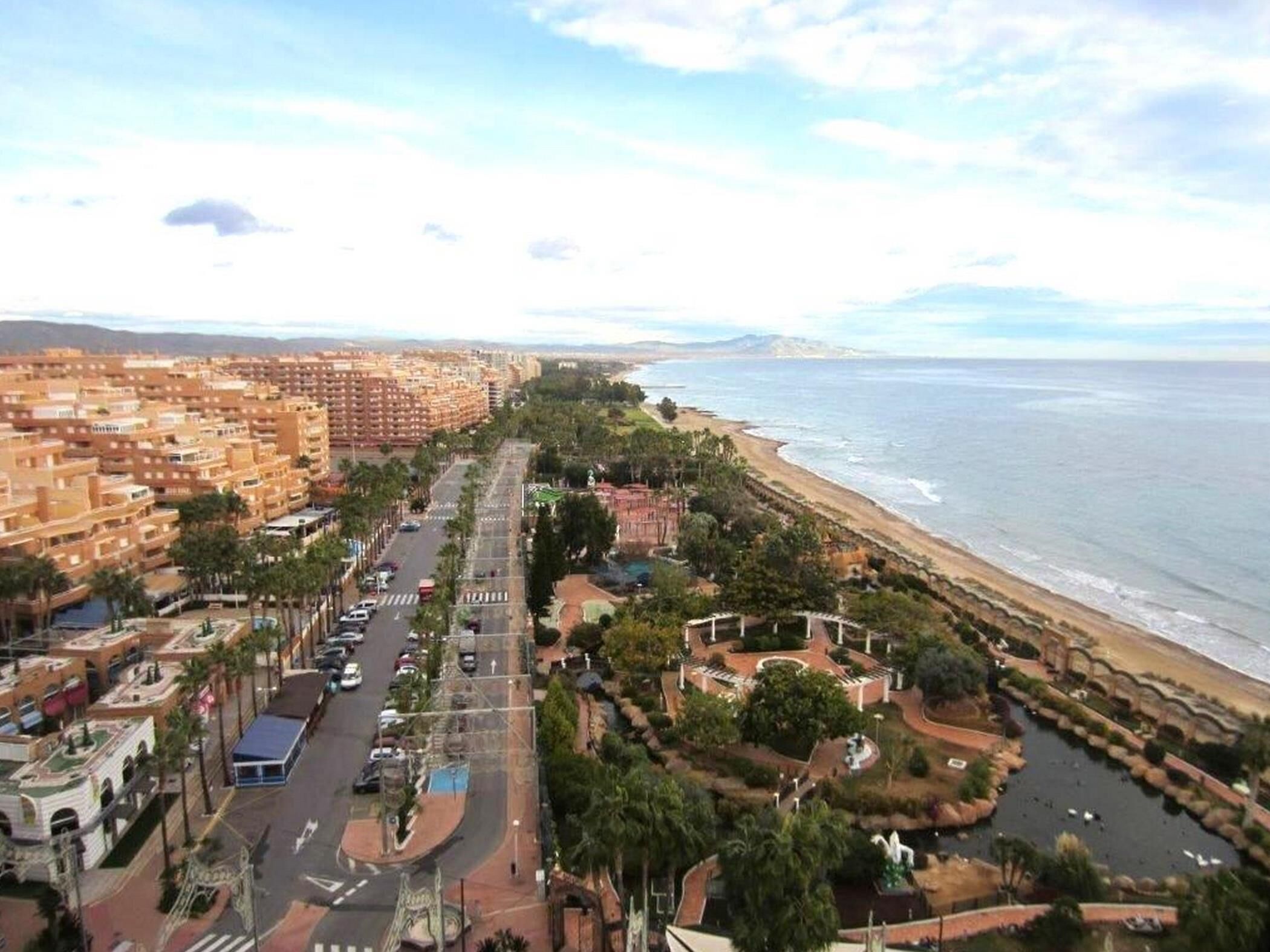 Un paysage urbain côtier avec une longue rue bordée d’immeubles, une plage et un parc agrémenté d’une fontaine.