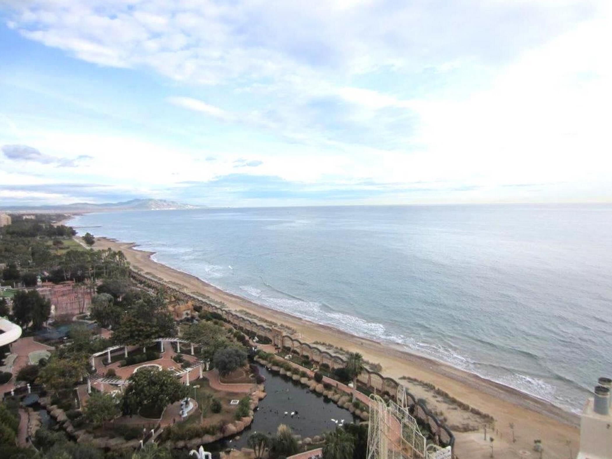A coastal view with a beach, a small water body, and a park area with pathways and greenery.