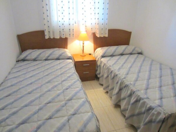 A room with two beds, each with a striped bedspread, a wooden headboard, and a bedside table with a lamp.