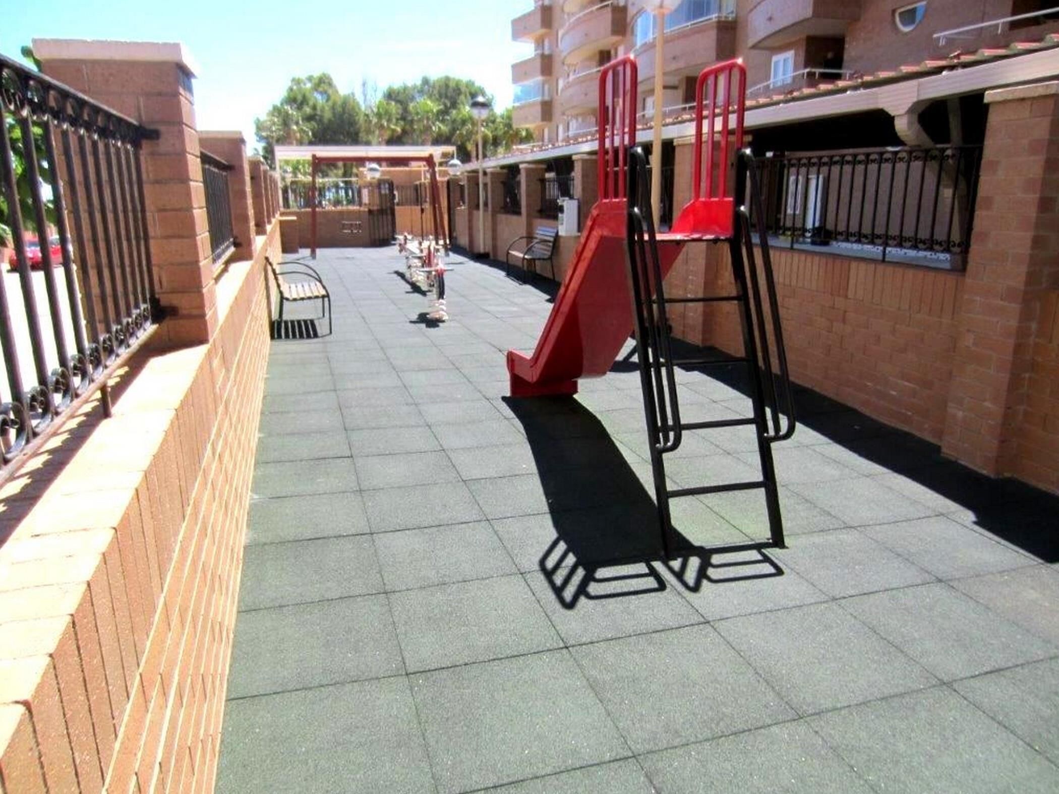 A playground area with a red slide and climbing structures, surrounded by brick walls and a metal fence.