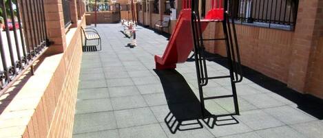 Zona de juegos infantil con un tobogán rojo y estructuras para escalar, rodeada de muros de ladrillo y una valla de metal.