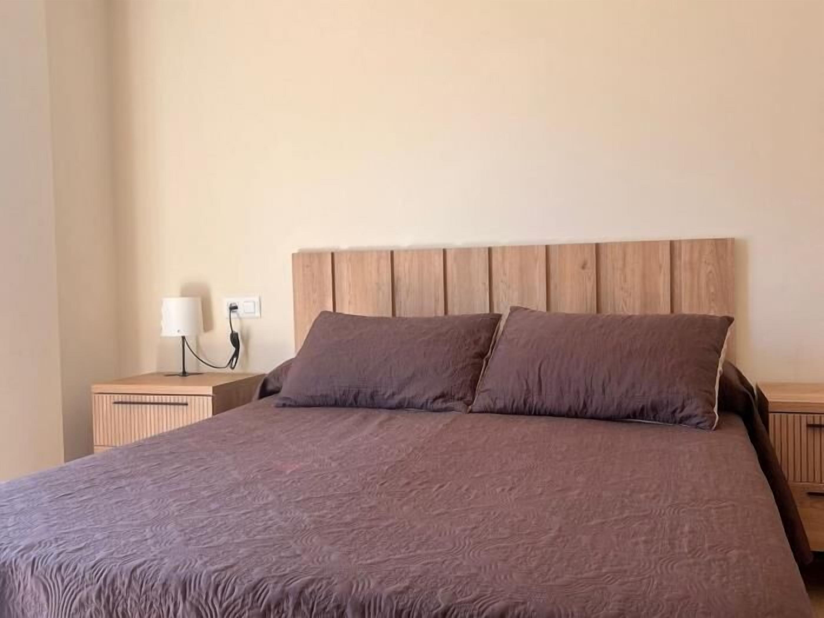 Un lit bien fait, avec une tête de lit en bois, une lampe de chevet et une prise électrique sur le mur.
