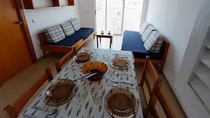 A compact living space with a dining table set for four, a sofa with blue cushions, a small table with a cup and saucer, and a view of the outside through a sliding door. - APCOSTAS Lotus / One Bedroom (Blanes)
