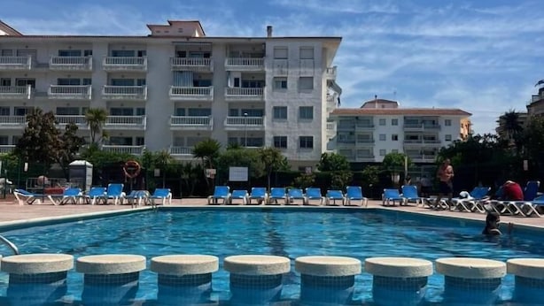 A swimming pool with blue water, surrounded by lounge chairs and a building with multiple balconies in the background. - APCOSTAS Lotus / One Bedroom (Blanes)