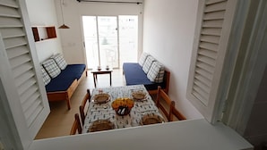 A compact dining area with a table set for four, two benches with cushions, a hanging pendant light, and a view through a sliding door to the outside. - APCOSTAS Lotus / One Bedroom (Blanes)