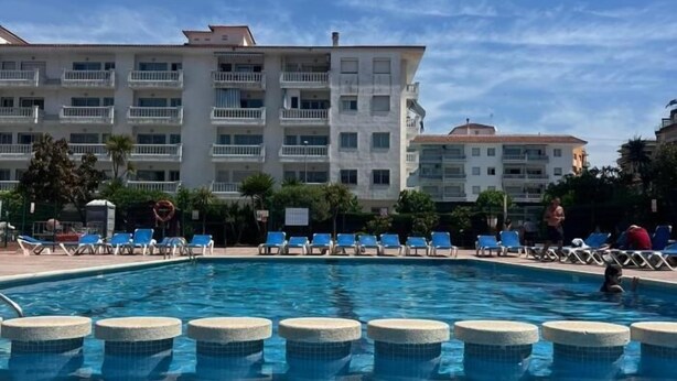 Una piscina con agua azul, rodeada de sillones de descanso y un edificio con varios balcones al fondo.