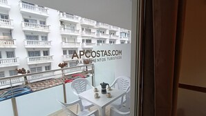 Un balcón con sillas y una mesa blancos, vista de un edificio blanco con balcones, y un cartel que dice "APCOSTAS.COM PARTEMENTOS TURISTICOS".