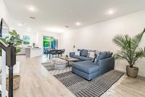 A modern living room with a blue sofa, a dining area with a table and chairs, and a kitchen area with a countertop and cabinets. - Luxury New Home in University Park Sarasota (Sarasota)