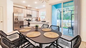Una cocina moderna con electrodomésticos de acero inoxidable, un comedor con una mesa oscura y individuales tejidos, y un amplio ventanal corredizo que da a un patio exterior.