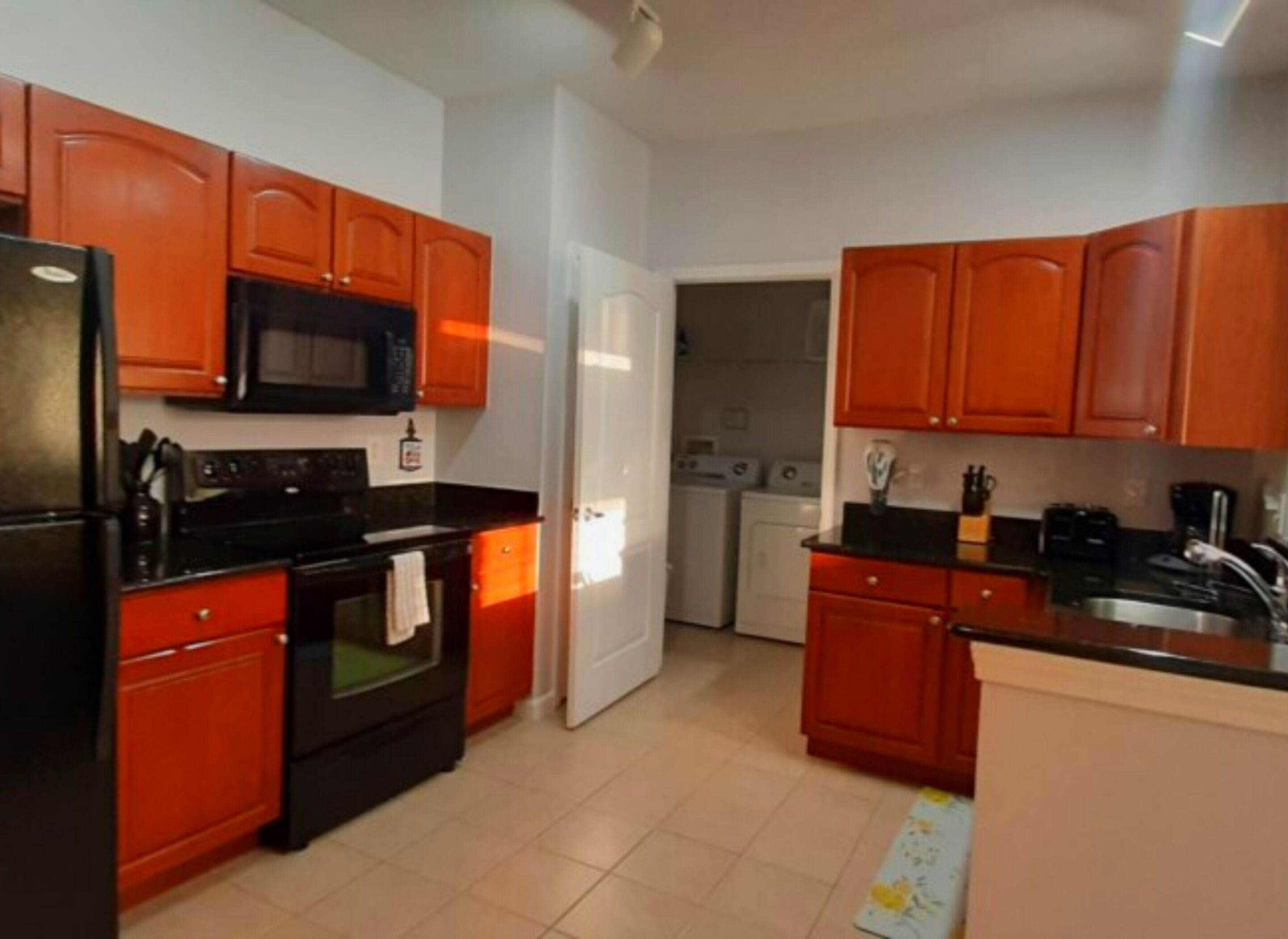 A compact kitchen with wooden cabinets, a black refrigerator, a microwave, a stove, and a washing machine in the adjacent room.