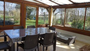 Een oranjerie met een glazen tafel en stoelen, grote ramen en uitzicht op een tuin.