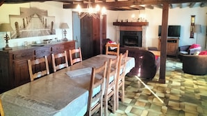 Een eetgedeelte met een grote tafel, houten stoelen, een kroonluchter, een open haard en een televisietoestel.