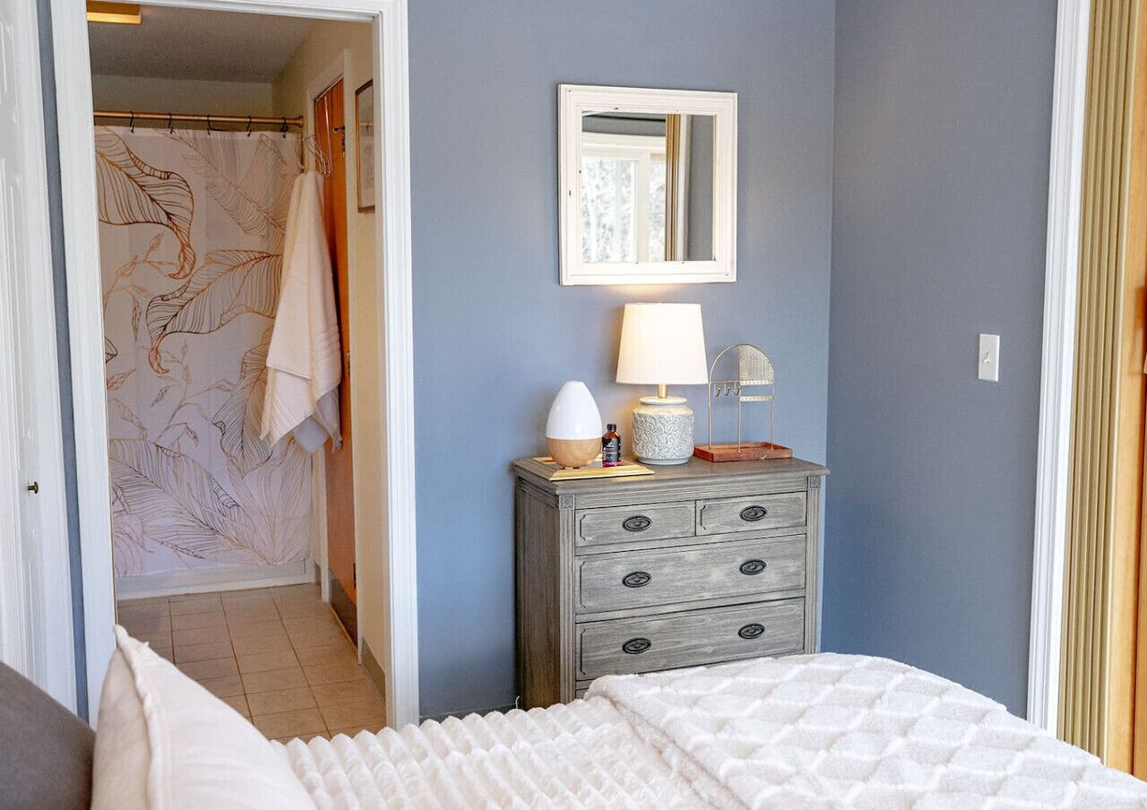 Une chambre à coucher avec un lit, une commode surmontée d’une lampe, un miroir et une douche dotée d’un rideau à motifs de feuilles.