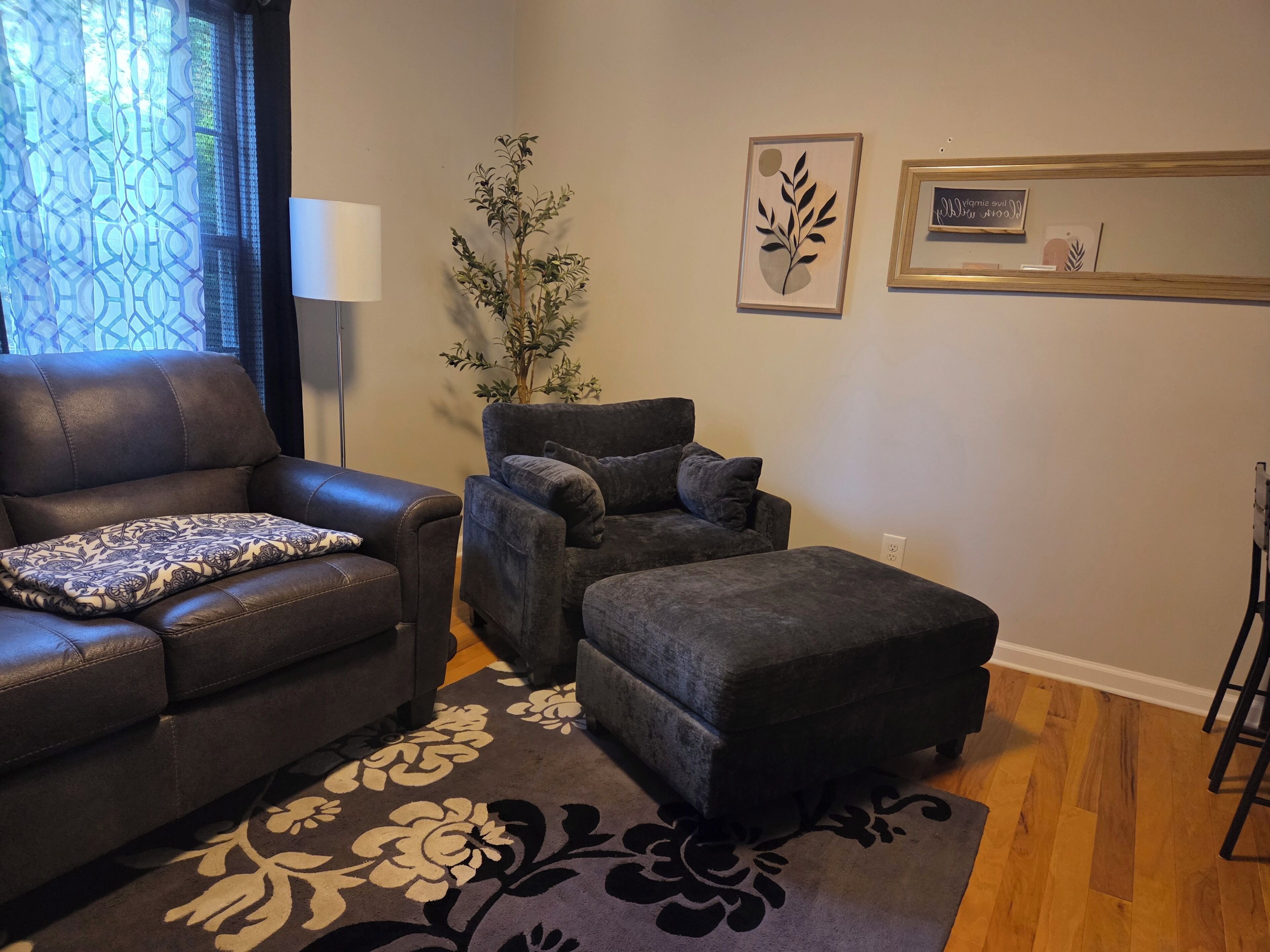 Un salon avec un canapé en cuir, un fauteuil gris et un pouf assorti. On y trouve un tapis à motifs sur le sol, une plante en pot et des œuvres d’art accrochées au mur.