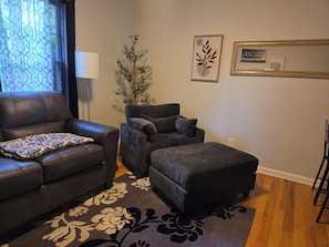 A living room with a leather sofa, a grey armchair, and a matching ottoman. There is a patterned rug on the floor, a potted plant, and wall art.