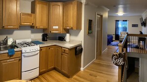 A kitchen with wooden cabinets, a stove, and a coffee maker. There is a hallway leading to a living area with a couch and a staircase.