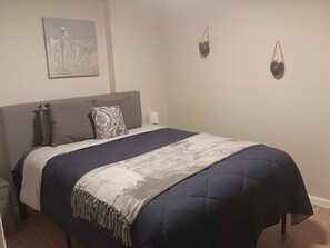 Una cama bien tendida con cabecera, una lámpara de noche y una obra de arte en la pared.