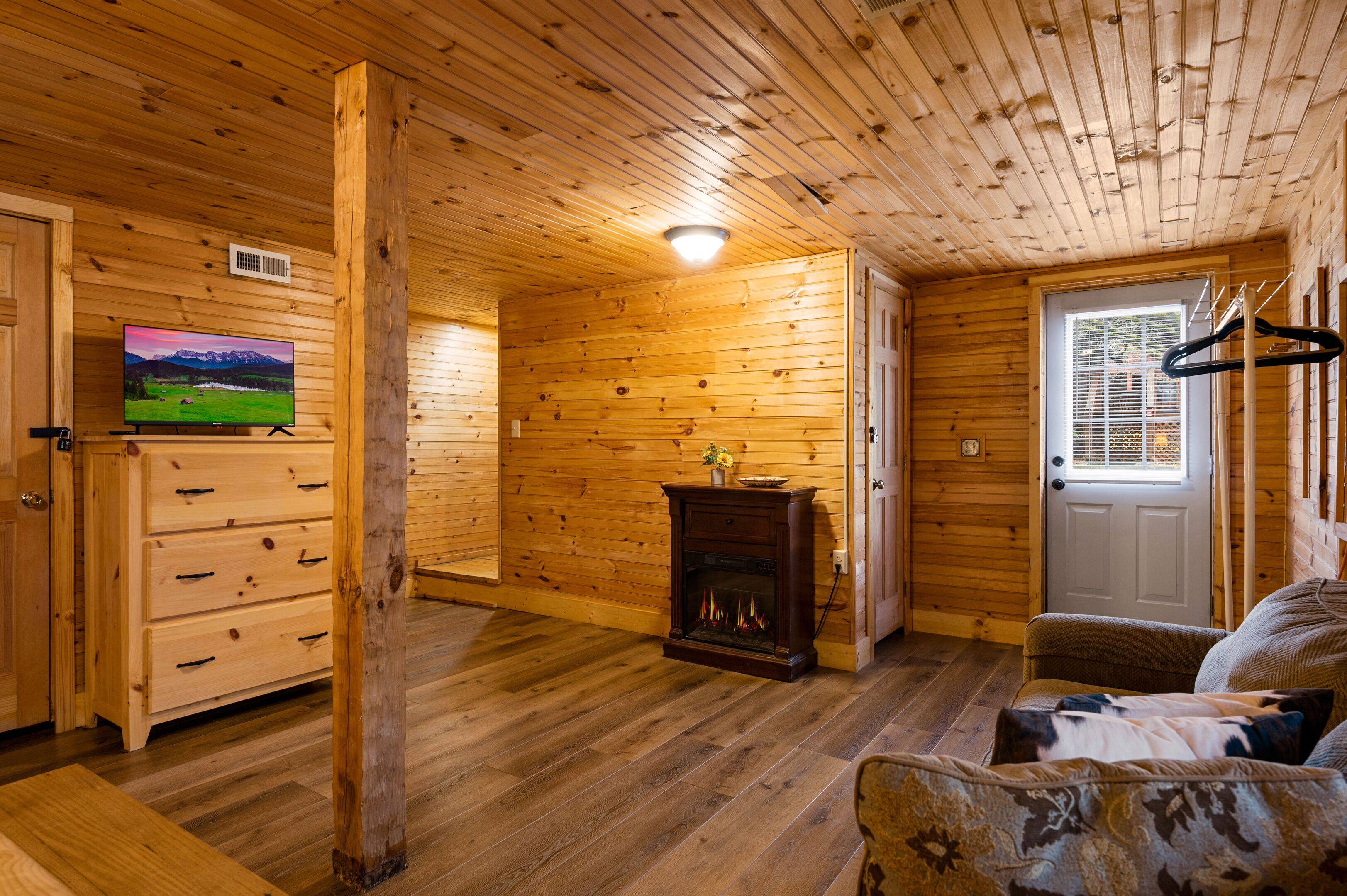 Interior de una cabaña de madera con chimenea, paredes de madera, un televisor y una puerta con ventanal.