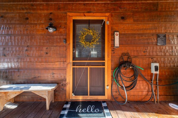 Entrada a una cabaña de madera con un banco, un adorno en la puerta y un felpudo que dice 'hola'.
